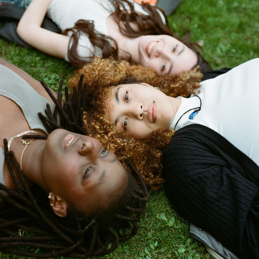 Three young women lying on the grass in a park. The woman in the center has curly hair, the woman on the left has dreadlocks and dark skin, and the woman on the right has straight hair. The group is relaxing outdoors.