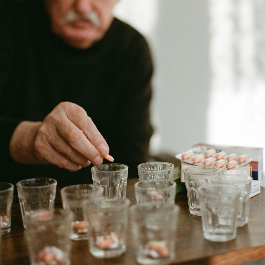 An elderly man, slightly blurred, wearing a black sweater, preparing medication by placing pills into glasses of water on a wooden table. Several pills and blister packs are visible.