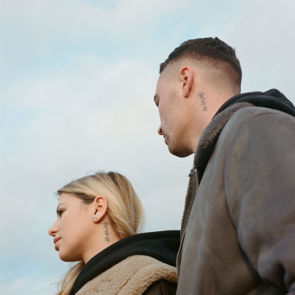 A couple seen from the side outdoors, both with Japanese tattoos on their necks. The woman has blonde hair, and the man has a short, shaved haircut. Both are wearing casual jackets on a cloudy day.