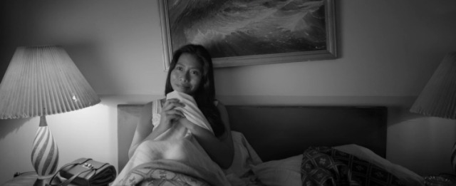 A woman sitting on a bed in a softly lit room, looking thoughtfully off-camera. The dim lighting creates an intimate and reflective atmosphere.