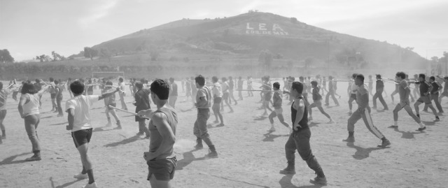 group of children running outdoors, possibly in a courtyard or playground, with hills visible in the background. The scene conveys energy and movement.