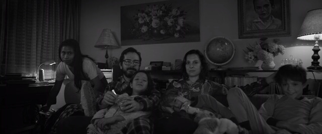 A family gathered on the couch, likely watching television together. The environment feels cozy, with homey decor details.