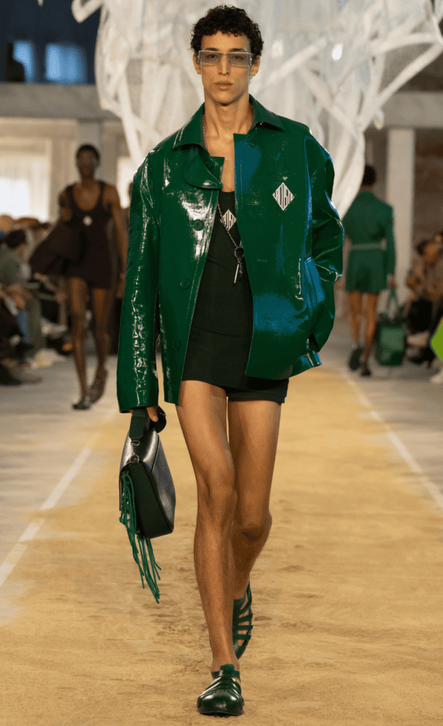 Runway image of a model wearing a glossy green jacket, black mini shorts, and matching accessories, representing modern minimalism in Spring/Summer 2025 fashion.