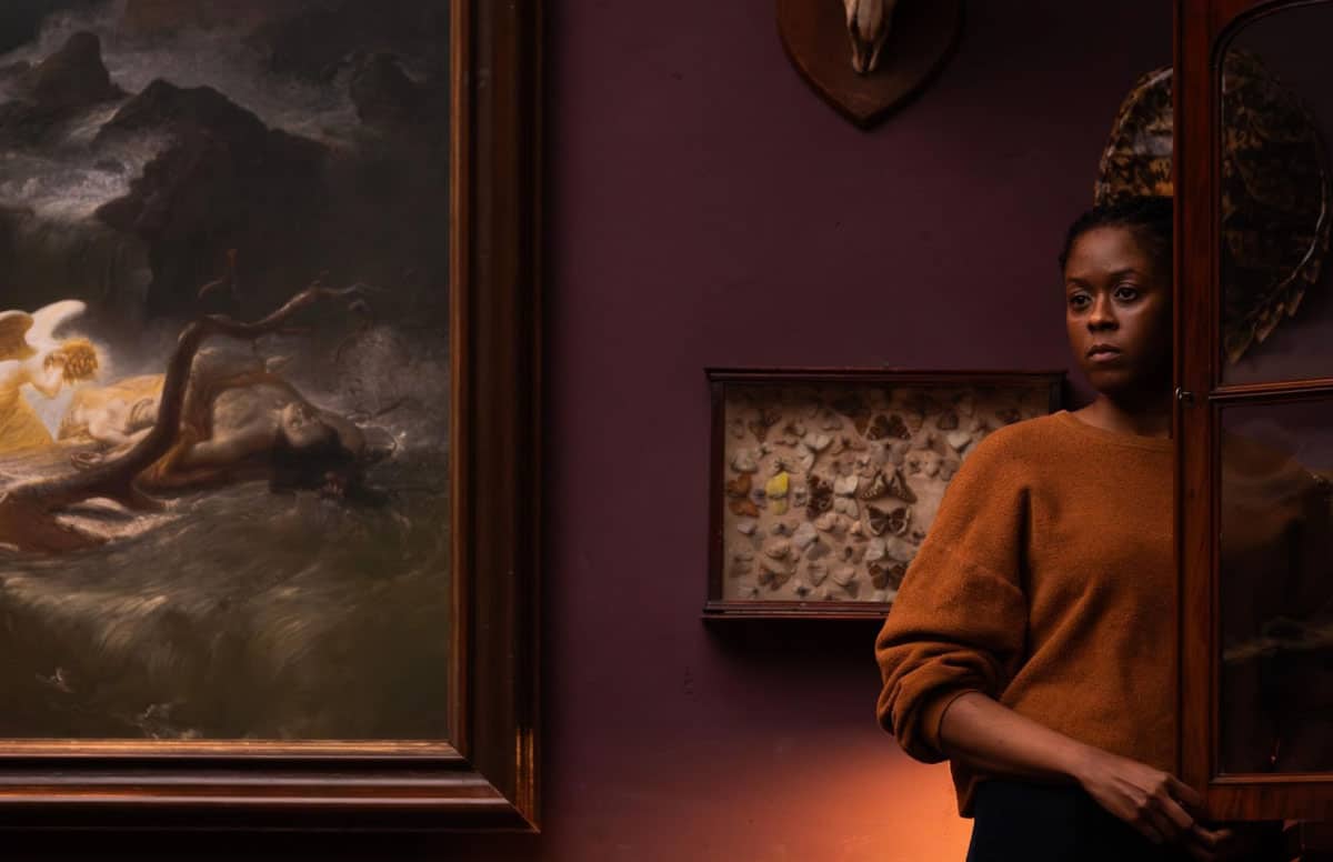 A woman standing in a dimly lit room beside a dramatic painting, evoking the eerie atmosphere of Joshua Oppenheimer’s film The End.