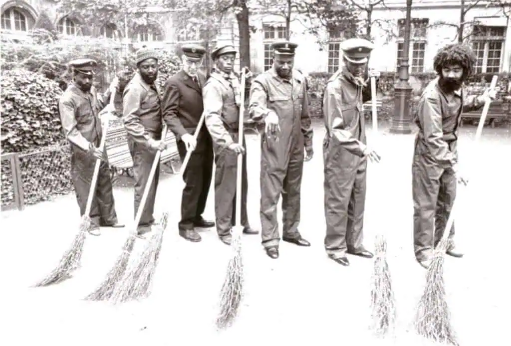Archival black-and-white image from Un Dessert Pour Constance (1980) showing seven men with brooms in formation, symbolising unity and postcolonial labour resistance.