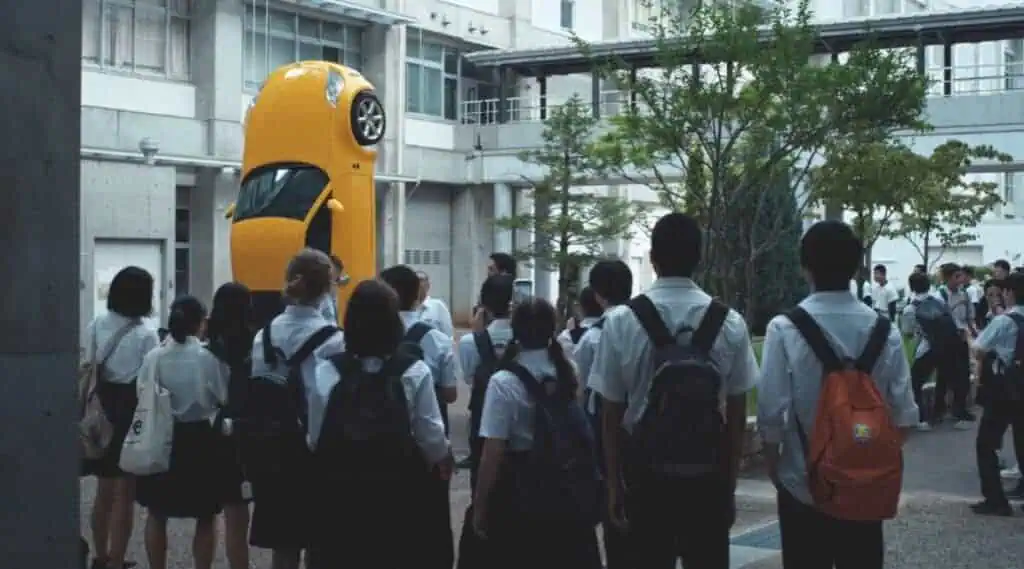 Still from Happyend (2024) showing students gathered around an upright yellow car installation in a school courtyard, representing absurdity and youth disillusionment.