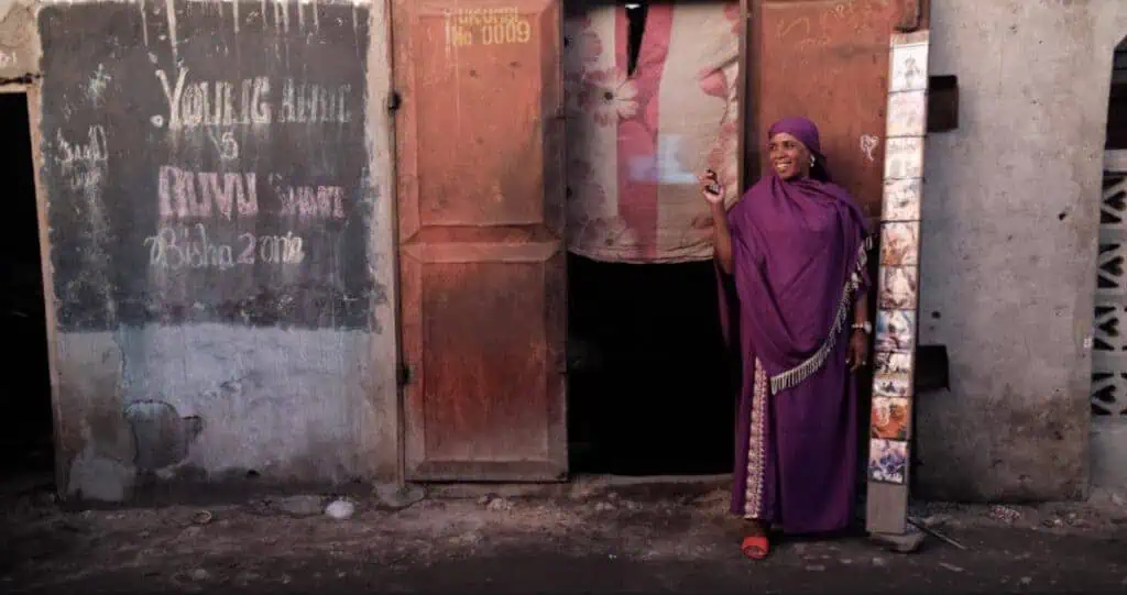 Scene from Apostles of Cinema (2023) showing a woman in a purple dress smiling outside a weathered building, embodying joy and community storytelling in East Africa.