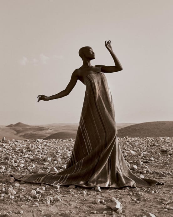 A Black woman in an artisanal dress with one hand up and one down