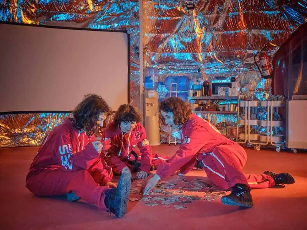 Three doppelgängers in red jumpsuits solving a puzzle in simulated space base interior