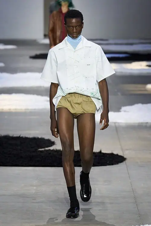 Model in a white short-sleeve shirt layered over a pale blue turtleneck and tan briefs, walking the runway in black loafers
