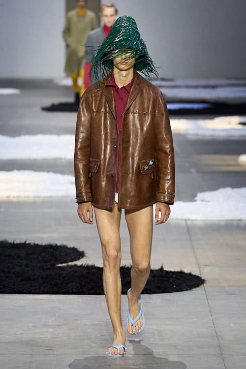 Model wearing a burgundy shirt under a brown leather jacket, styled with flip-flops and a green shredded hat obscuring the face