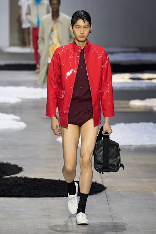 Model wearing a glossy red jacket over a burgundy shirt and high-cut briefs, paired with white sneakers and carrying a black duffle bag