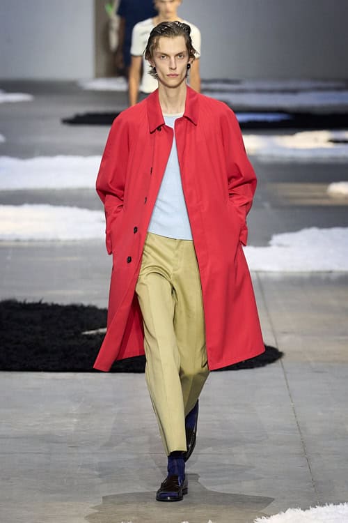 Model in a red trench coat over a pale blue sweater and beige trousers, styled with blue socks and black loafers