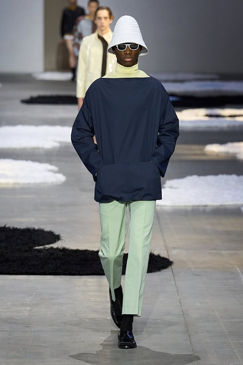 Model wearing a white bucket hat, black sunglasses, and navy pullover with mint green trousers, walking confidently down the runway
