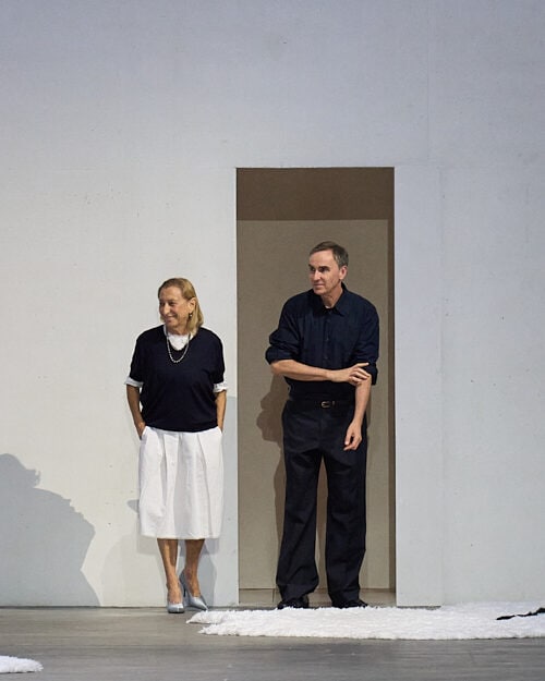 Two designers standing in a minimal doorway at the end of a fashion show runway, acknowledging the audience after the show