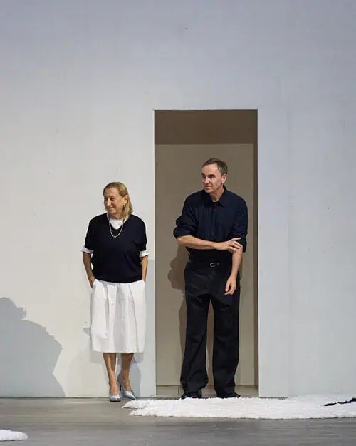 Two designers standing in a minimal doorway at the end of a fashion show runway, acknowledging the audience after the show