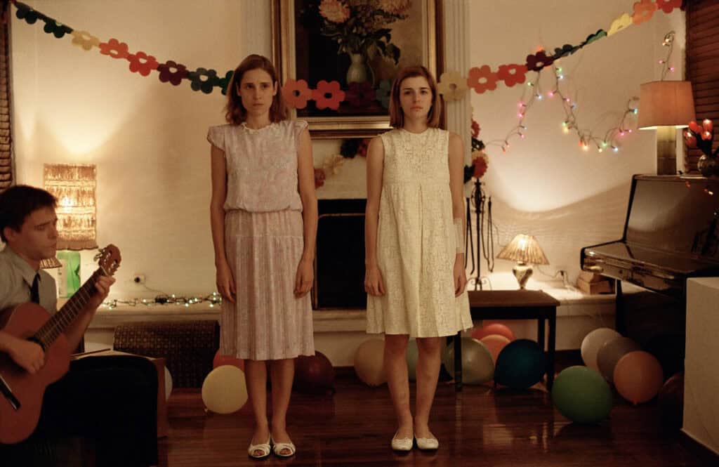 Two sisters stand motionless in pastel dresses during a tense birthday party, with decorations and a guitar player in the background – scene from Dogtooth (2009)
