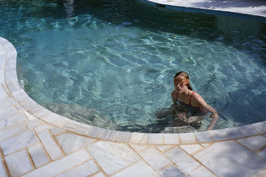 A blindfolded young woman sits partially submerged in a pool, emphasizing the absurd control and isolation of the children in Dogtooth (2009)