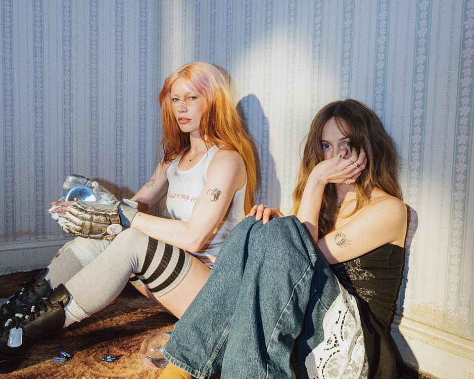 Two women sit against vintage wallpaper, one with long red hair and hockey gloves holding a chrome orb, the other peeking from behind her hand, creating a dreamlike, eerie mood.