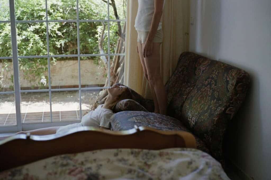 A girl lies on a couch while another stands beside her, framed by a large window and overgrown garden, capturing the eerie domesticity of Dogtooth (2009)