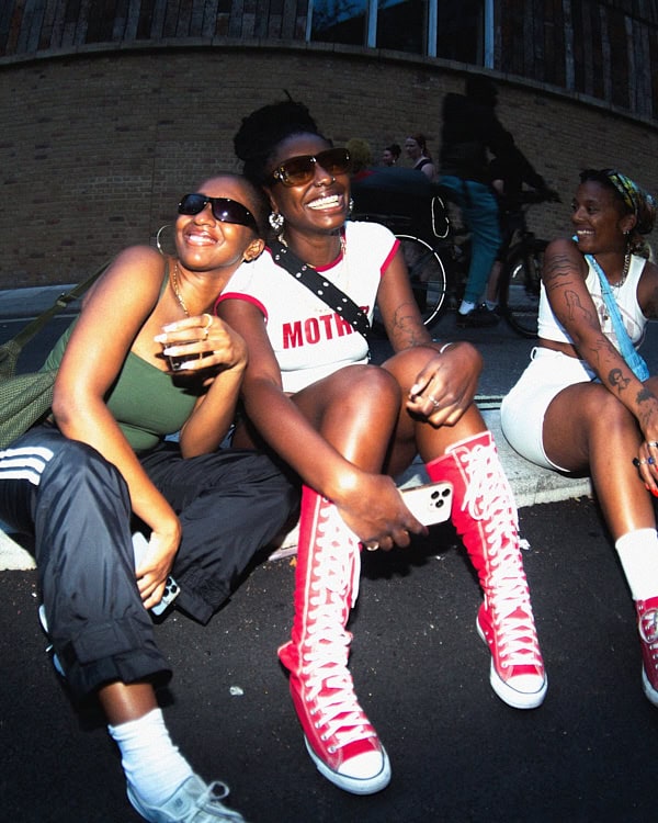 Three women sitting on a London street in streetwear and high-top Converse, smiling and enjoying A Voice Amplified