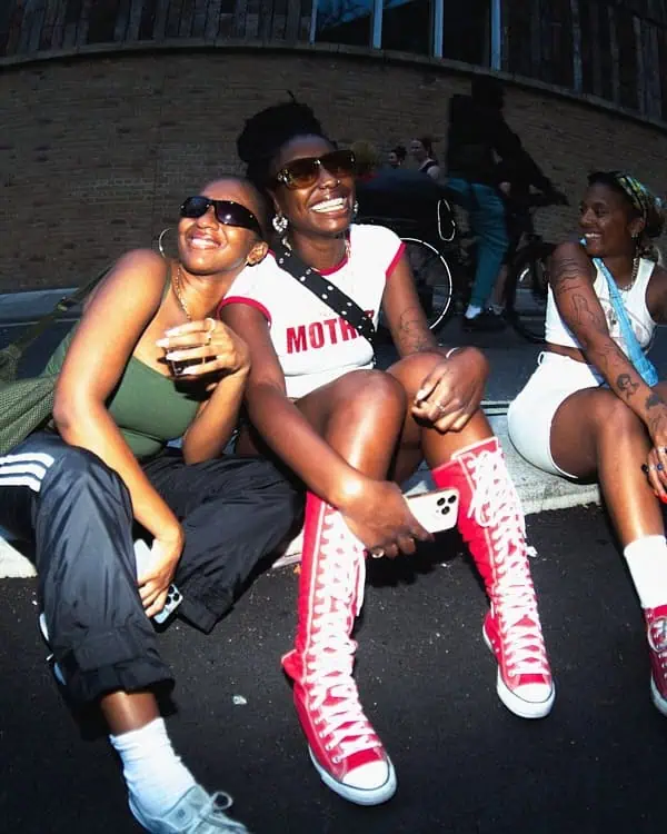 Three women sitting on a London street in streetwear and high-top Converse, smiling and enjoying A Voice Amplified