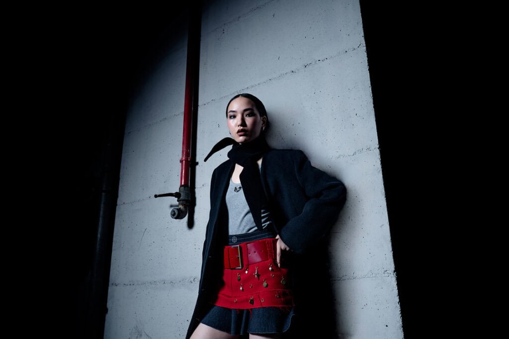 Model posing against a concrete wall with a red pipe, wearing a black coat and a red embellished belt