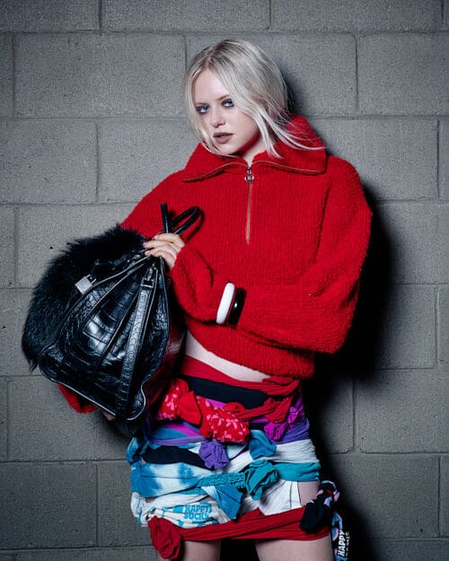 Blonde model in a red zip-up sweater, holding a black leather bag, with a skirt made of colorful knotted fabrics