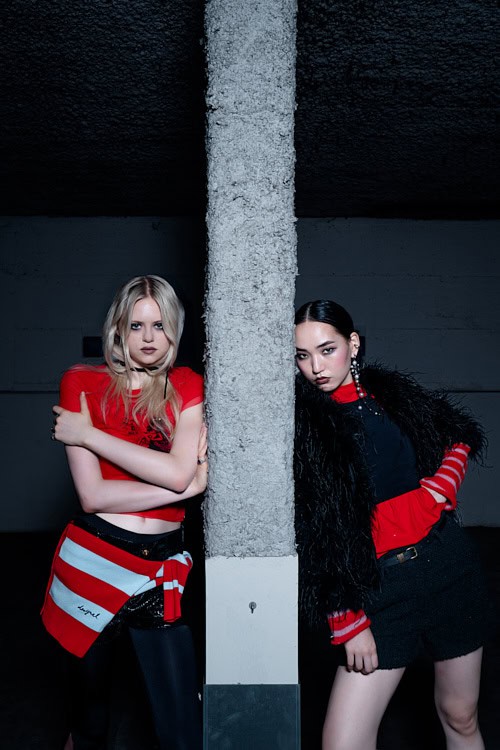 wo models leaning against a pillar in a parking garage, wearing red and black contrasting outfits
