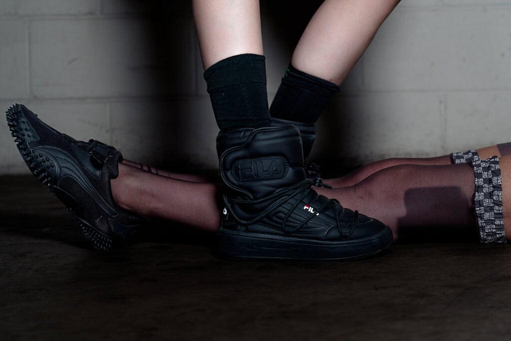 Close-up of intertwined legs wearing black Fila and Puma shoes, against a concrete background