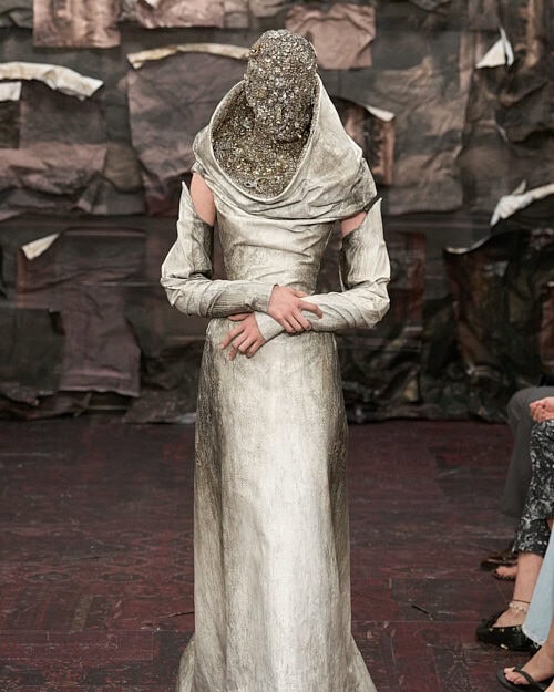 Runway model wearing a sculptural, metallic-toned gown with a beaded, full-face headpiece during a Margiela fashion show