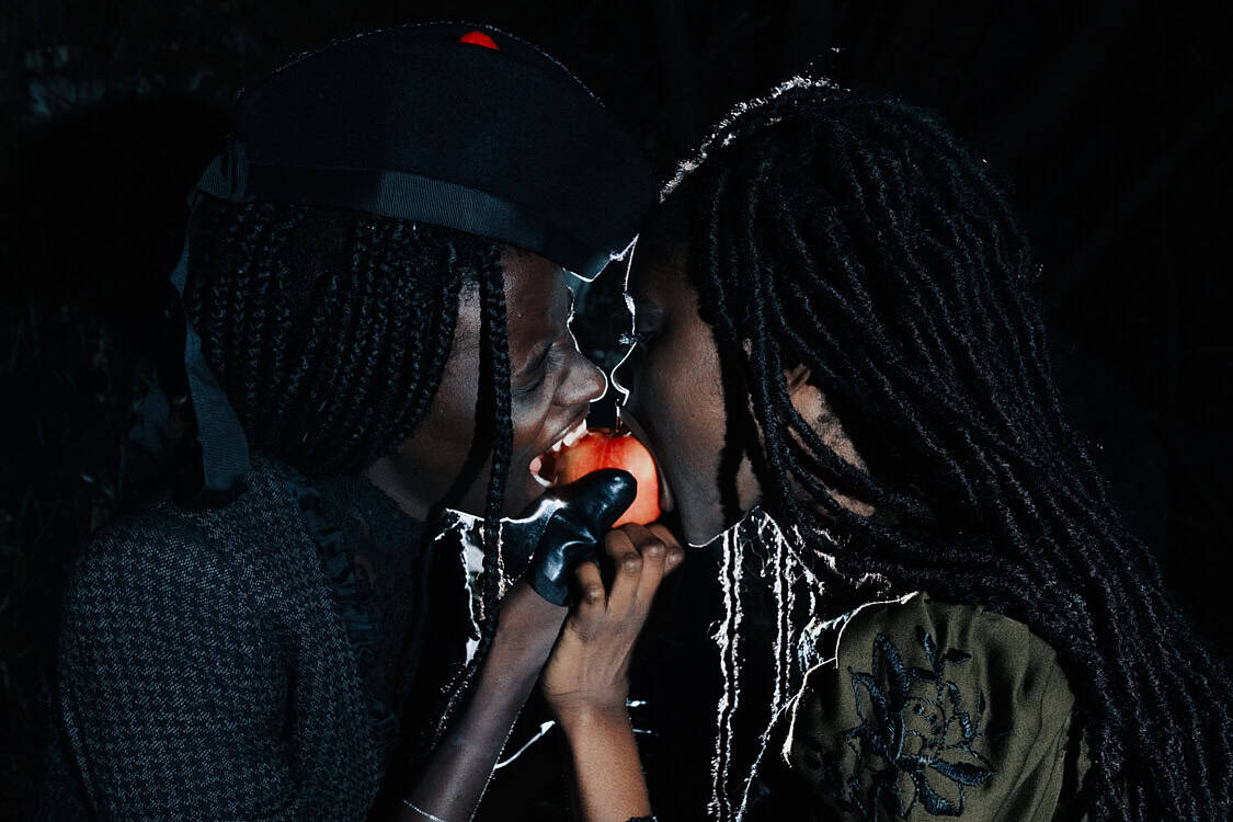 Blonde model in a red zip-up sweater, holding a black leather bag, with a skirt made of colorful knotted fabrics