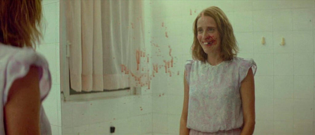 A young woman smiles at her own reflection in a blood-smeared bathroom mirror, evoking the disturbing consequences of psychological manipulation – Dogtooth (2009)