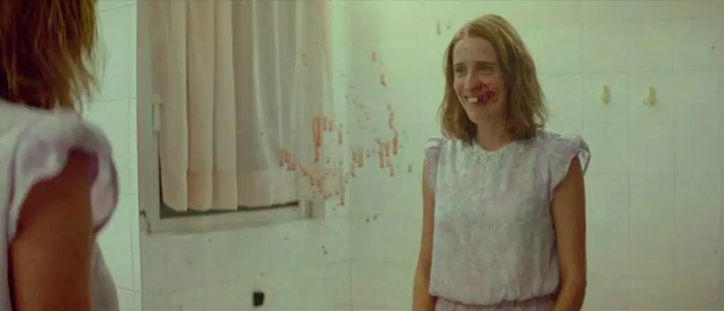 A young woman smiles at her own reflection in a blood-smeared bathroom mirror, evoking the disturbing consequences of psychological manipulation – Dogtooth (2009)