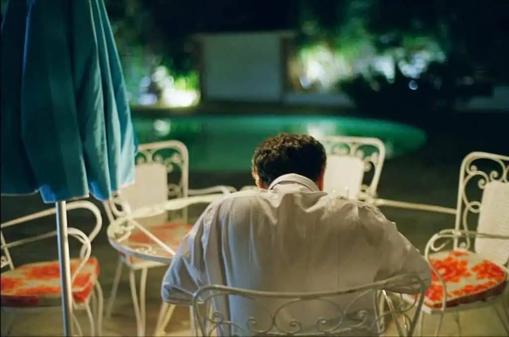 The father sits alone at a table by the pool at night, his back turned, reflecting the oppressive control and alienation in Dogtooth (2009)