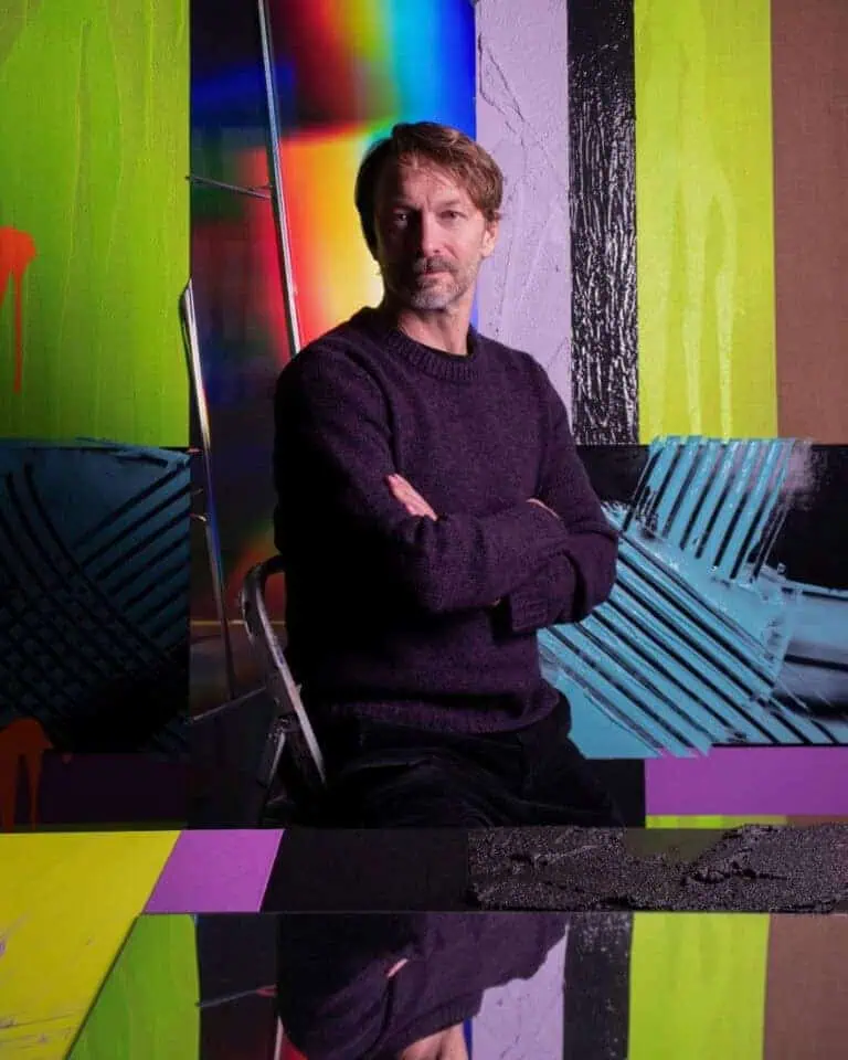 Portrait of an artist seated with arms crossed in front of colorful abstract paintings featuring vivid green, purple, and black textures.