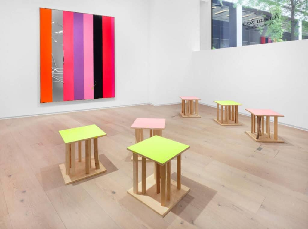 Minimalist gallery installation with neon-colored wooden stools and a striped artwork on the wall, combining vibrant pinks, oranges, and metallic elements.