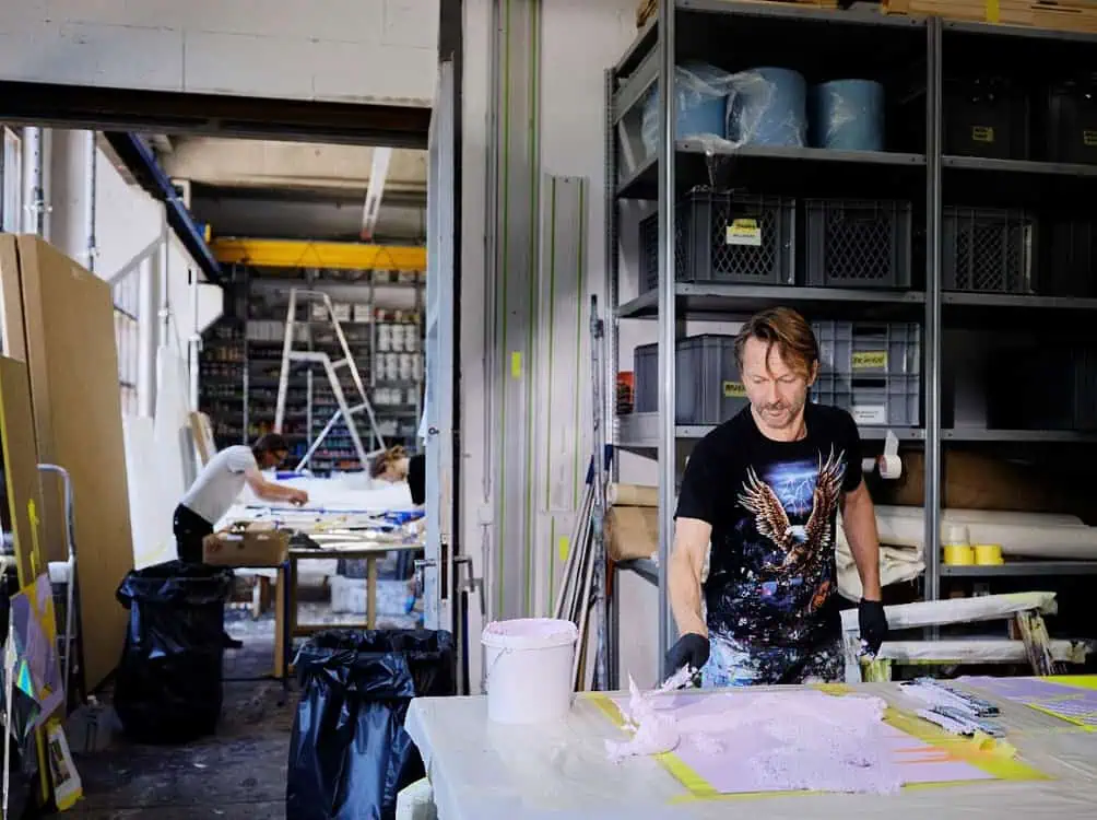 Artist working with pink material in a studio filled with tools, supplies, and shelves, while others work in the background.