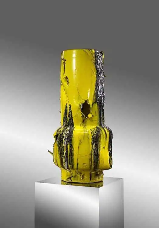 Yellow sculptural object resembling a compressed metal container, partially coated with black paint and displayed on a pedestal.