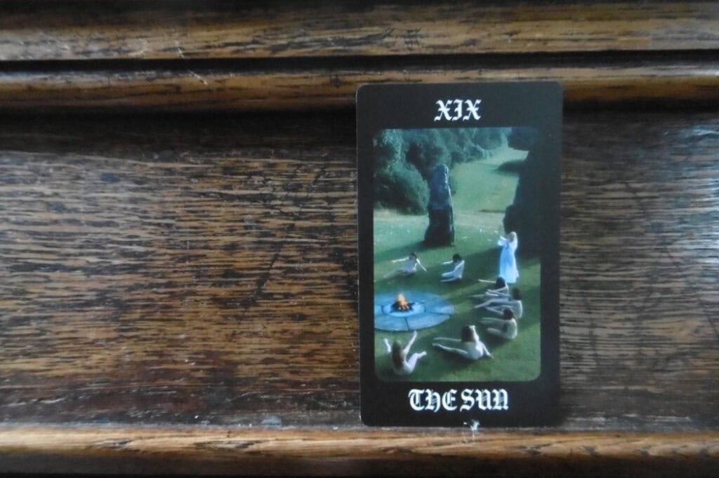 Tarot card titled 'The Sun', depicting a ritual scene with nude figures sitting in a circle around a small fire on a grassy field.