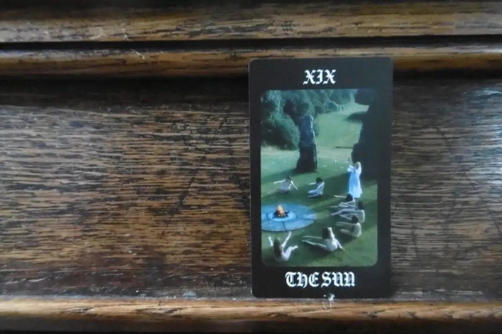 Tarot card titled 'The Sun', depicting a ritual scene with nude figures sitting in a circle around a small fire on a grassy field.