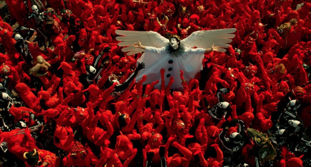 A central figure dressed as a white angel with wings outstretched, held aloft by a dense crowd of people in red costumes and masks, creating a surreal and theatrical scene.