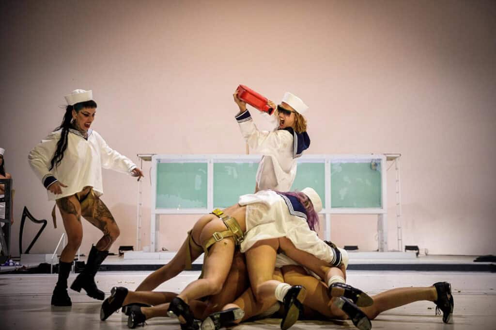 Group of performers in sailor outfits creating a human pile, while another sprays red liquid from a canister