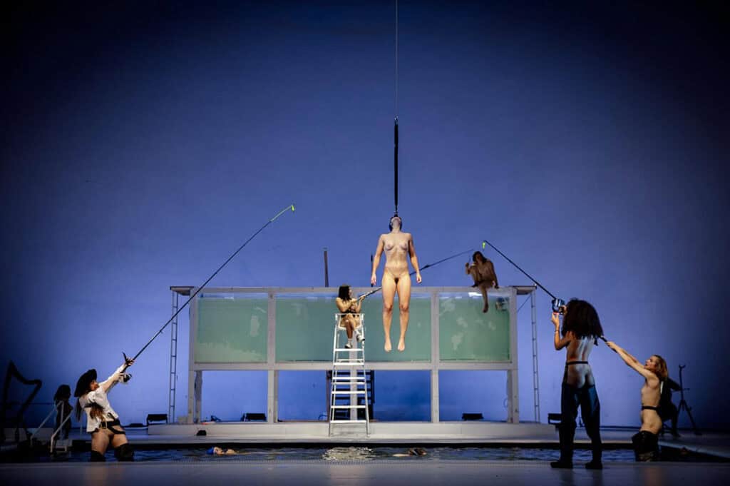 Nude performer suspended by a hook above a transparent tank, surrounded by others using fishing rods on stage