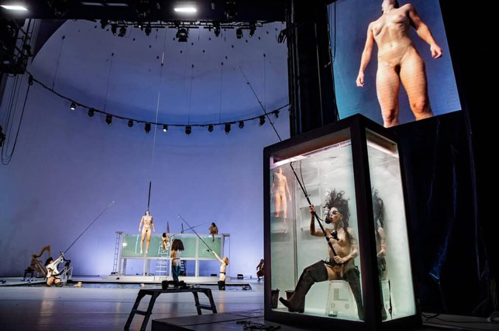 Theatrical stage with multiple performers interacting with water tanks and fishing rods, spotlight on a diver inside a tank