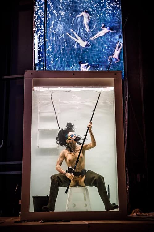Performer submerged in a transparent water tank, wearing a diving mask and holding a fishing rod, with a projection of swimmers above