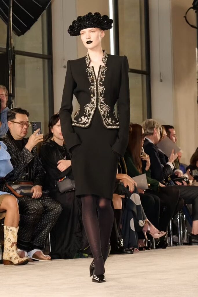 Model in a structured black suit jacket with ornate metallic embroidery, paired with a black sculptural hat and dramatic black lipstick