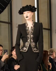 Model in a structured black suit jacket with ornate metallic embroidery, paired with a black sculptural hat and dramatic black lipstick