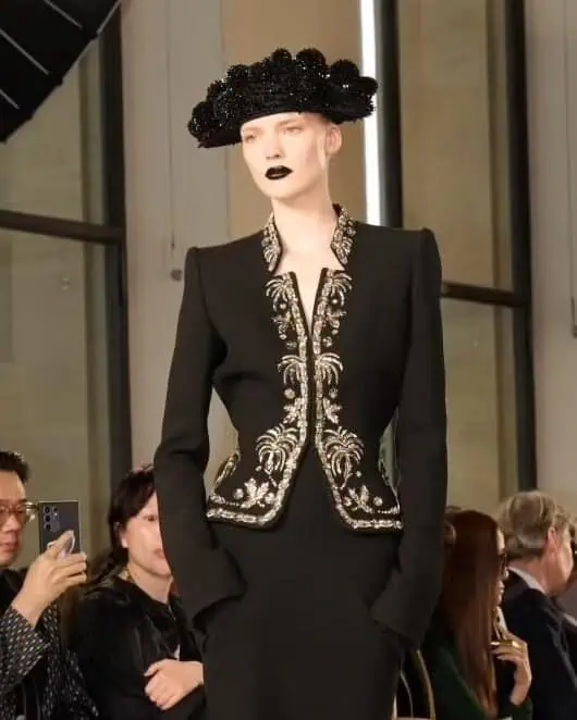 Model in a structured black suit jacket with ornate metallic embroidery, paired with a black sculptural hat and dramatic black lipstick