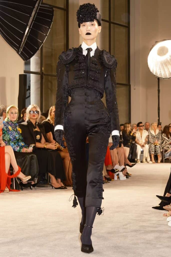 Model wearing a black embellished matador-style suit with exaggerated hip structure, black gloves, and dramatic headpiece on the runway